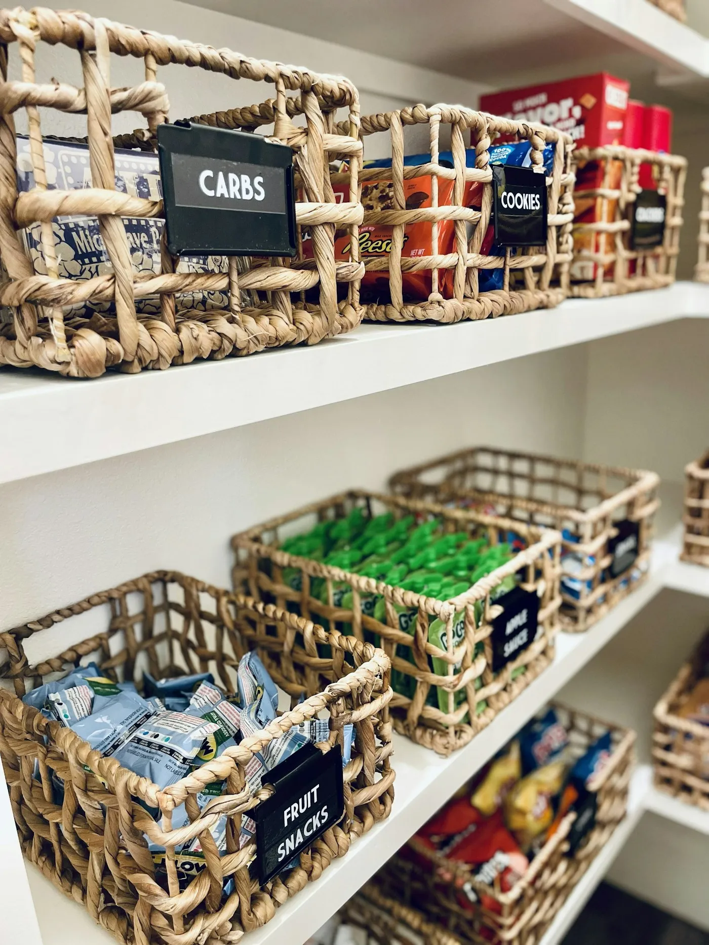 A bright pantry with woven baskets on white shelves, each labeled: carbs, cookies, fruit snacks, apple sauce.